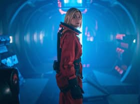 Jodie Whittaker as the Doctor, wearing an orange Sanctuary Base 6 spacesuit, turning back to look at the camera in a blue-toned spaceship corridor (Credit: James Pardon/BBC Studios)