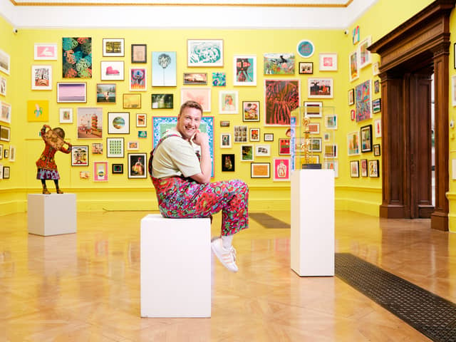 Comedian Joe Lycett, wearing floral dungarees and smiling, sat on a white art display plinth in the Royal Academy. The walls are yellow and covered in framed paintings (Credit: BBC Studios/Richard Ansett)