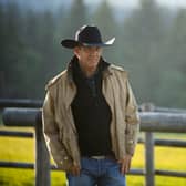 Kevin Costner as John Dutton in Yellowstone, stood in a field next to a fence, his hands in his pockets, wearing a cowboy hat (Credit: Paramount+) 