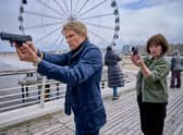 Marc Warren as Van der Valk as Maimie McCoy as Lucienne, holding their guns straight ahead. They’re on a pier, with a ferris wheel behind them. (Credit: ITV)