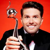 Joel Dommett, wearing a tuxedo in front of a block red background, holding out an NTA towards the viewer and smiling (Credit: ITV)