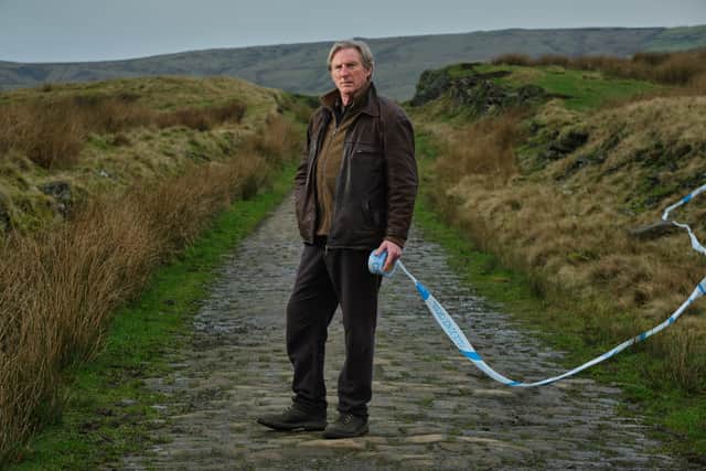 Adrian Dunbar as Alex Ridley, stood on a cobbled street running through a field. Police ticker tape floats on the wind behind him. (Credit: ITV)