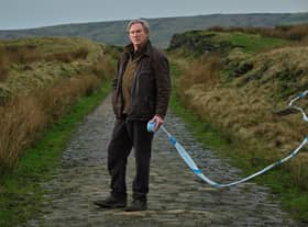 Adrian Dunbar as Alex Ridley, stood on a cobbled street running through a field. Police ticker tape floats on the wind behind him. (Credit: ITV)
