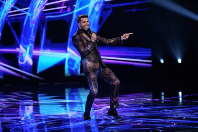 Joel Dommett gesturing wildly while hosting The Masked Dancer. He’s wearing a suit with a starfield pattern on it (Credit: ITV)
