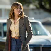 Ophelia Lovibond as Joyce in Minx. She’s wearing a yellow shirt and blue jeans underneath a checked coat; there’s a car behind her, out of focus in the background (Credit: Katrina Marcinowski/HBO Max)