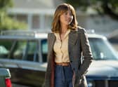 Ophelia Lovibond as Joyce in Minx. She’s wearing a yellow shirt and blue jeans underneath a checked coat; there’s a car behind her, out of focus in the background (Credit: Katrina Marcinowski/HBO Max)