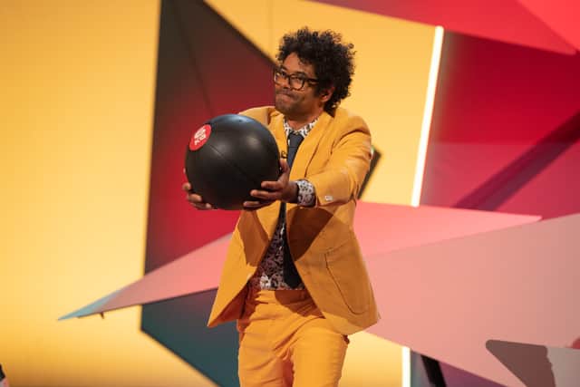 Richard Ayoade on Question Team, wearing a yellow suit and holding a large black sphere in outstretched hands. The sphere is labelled “6kg” (Credit: UKTV)