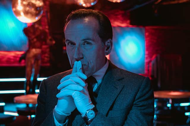 Ben Miller as Professor T, stood in a dark red-lit room, wearing surgical gloves, his hands steepled in front of him (Credit: Sofie Gheysens/ITV)