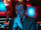 Ben Miller as Professor T, stood in a dark red-lit room, wearing surgical gloves, his hands steepled in front of him (Credit: Sofie Gheysens/ITV)
