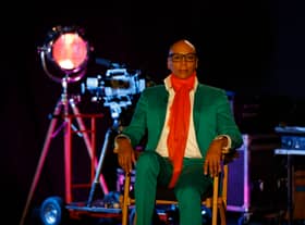RuPaul is a drag megastar wearing a green suit and orange scarf with a large lamp in the background