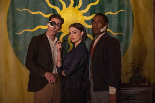 A still from Amsterdam (2022), depicting Christian Bale as Burt, Margot Robbie as Valerie, and John David Washington as Harold. Burt is wearing an eyepatch, Valerie is holding a smoking pipe aloft, and Harold has a bandage on his cheek; behind them is a yellow curtain with a golden sun in a green circle emblazoned across its centre (Credit: 20th Century Studios)