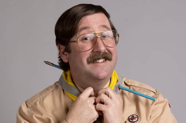 Jim Howick as scout leader Pat in Ghosts, adjusting his scarf. He’s got a mustache, big round glasses, and an arrow through his neck (Credit: BBC/Monumental/Guido Mandozzi) 