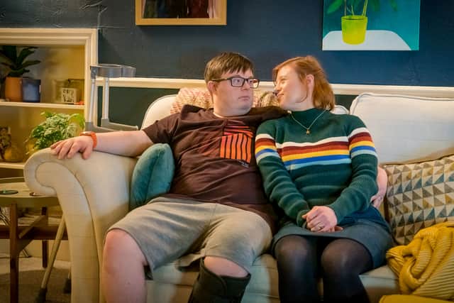Leon Harrop as Ralph and Sarah Gordy as Katie, sat together on the sofa. Ralph has his leg in a cast and there are crutches to one side of the sofa (Credit: ITV Studios/BBC)