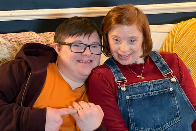 Leon Harrop as Ralph and Sarah Gordy as Katie in Ralph and Katie. They’re sat together on a sofa, leaning into one another and smiling. Ralph is gesturing at his wedding ring (Credit: BBC / ITV Studios)