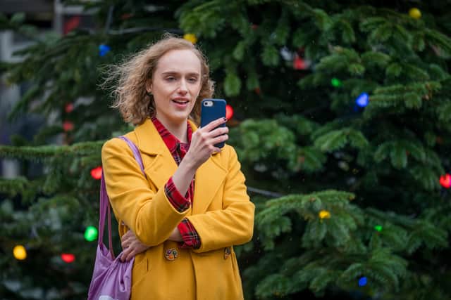 Matt Greenwood as Tom in Ralph & Katie, standing in front of a big outdoor Christmas tree. He’s wearing a yellow coat over a red shirt, holding his blue phone out in front of him (Credit: BBC/ITV Studios)