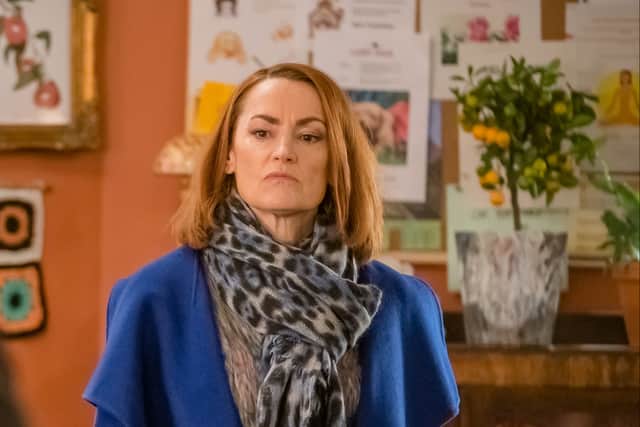 Pooky Quesnel as Louise in Ralph & Katie, looking frustrated. She’s wearing a blue coat and a grey scarf with black dots; behind her is the bakery notice board and a plant with yellow fruit. (Credit: BBC/ITV Studios)
