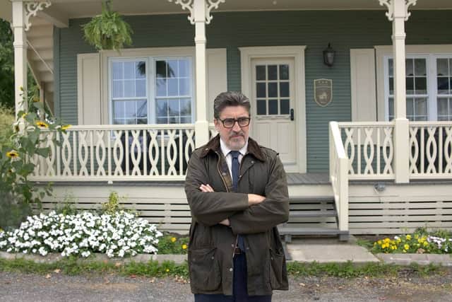 Alfred Molina as Armand Gamache in Three Pines, stood outside a house with his arms folded (Credit: Amazon Studios)