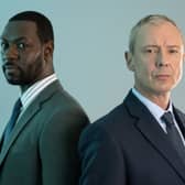 Richie Campbell as DS Glenn Branson and John Simm as DI Roy Grace in Grace S3, stood against a soft green background (Credit: ITV)