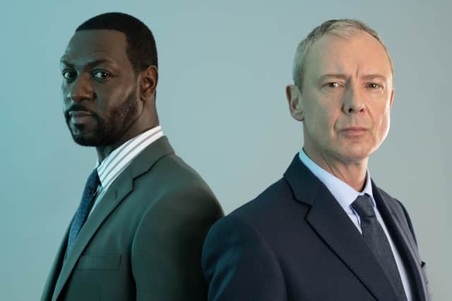 Richie Campbell as DS Glenn Branson and John Simm as DI Roy Grace in Grace S3, stood against a soft green background (Credit: ITV)