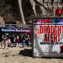 A couple uses a selfie stick to take a picture next to a banner warning tourists on drought alert in Catalonia, near Sagrada Familia basilica in Barcelona on April 13, 2024. From the Balearic Islands to the Canary Islands, Barcelona and Malaga, anti-mass tourism movements are multiplying in Spain. On the Canary Island Tenerife, activists started hunger strike on April 11 to demand a moratorium on mass tourism. (Photo by PAU BARRENA / AFP) (Photo by PAU BARRENA/AFP via Getty Images)